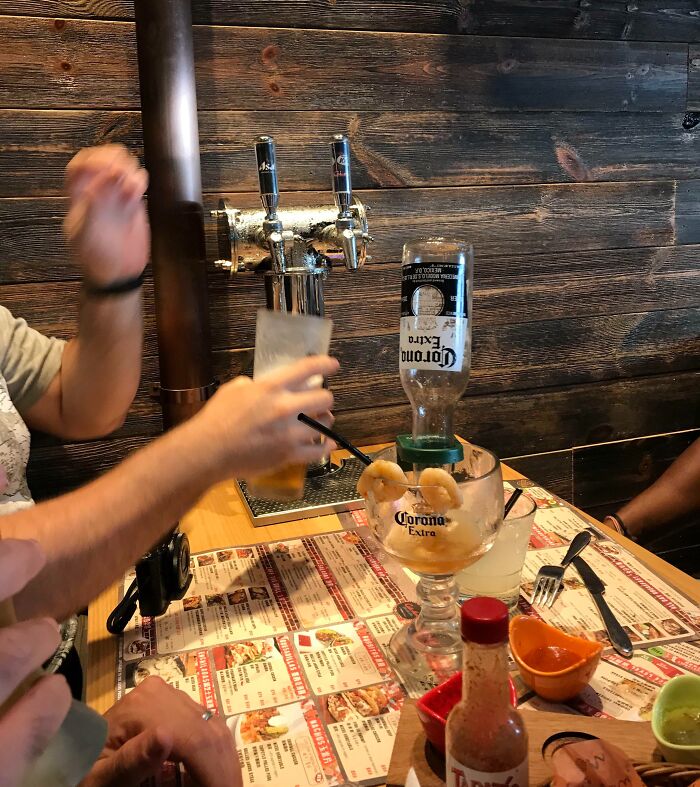 Creative bar setup with a Corona Extra bottle, beer tap, and drinks on a wooden table in a lively bar environment.