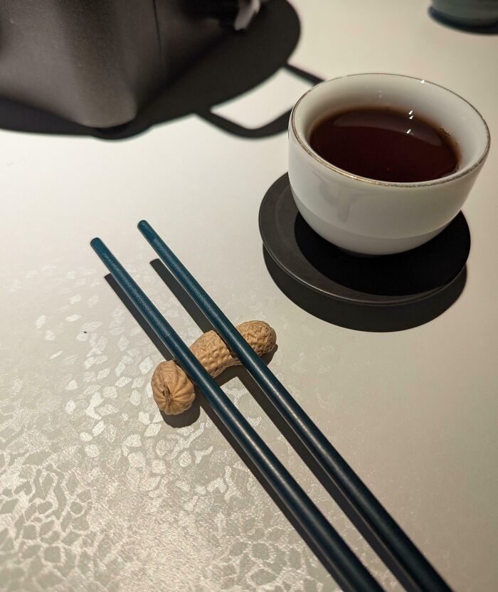 White ceramic cup with dark tea, black saucer, and blue chopsticks resting on peanuts at a creative bar or restaurant setting.