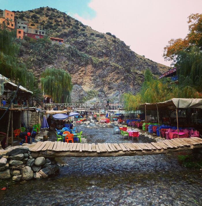 Open-air bars and restaurants set along a mountain stream with colorful chairs and wooden footbridges showcasing creativity.