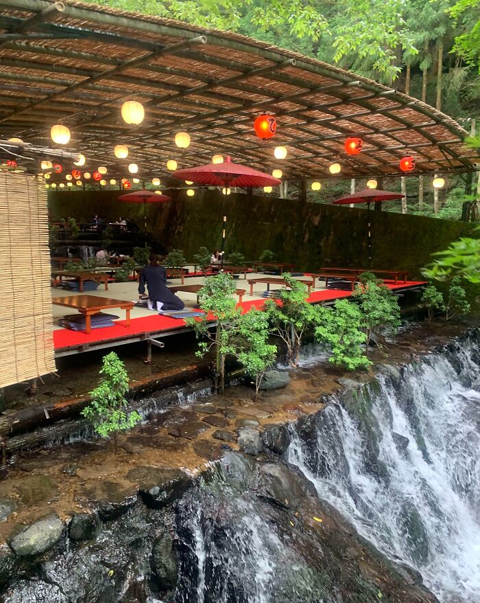 Outdoor bar and restaurant with red umbrellas and lanterns beside a flowing waterfall in a creative natural setting.