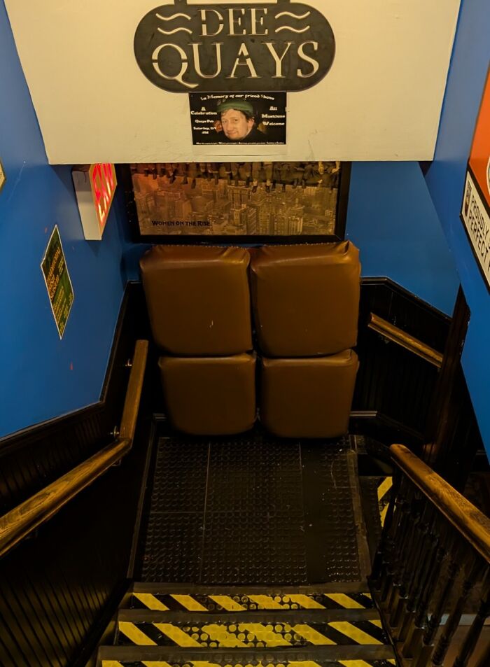Staircase entrance with brown seats blocking the way inside Dee Quays bar, featuring blue walls and wooden railings.