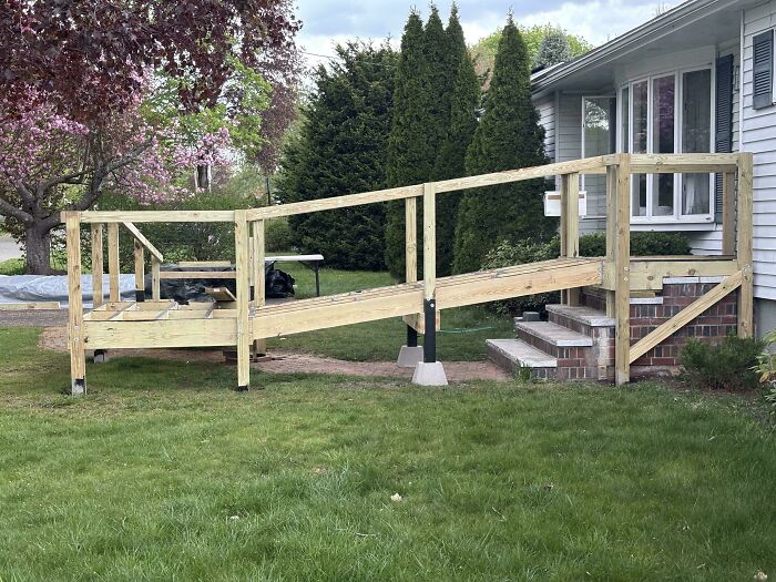 Wooden accessibility ramp under construction leading to a house porch, surrounded by green lawn and trees, wholesome kind neighbors.