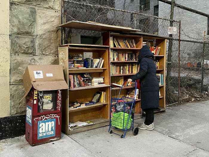 Person in a black coat browsing an outdoor free library, showcasing wholesome kind neighbors sharing books on the street.