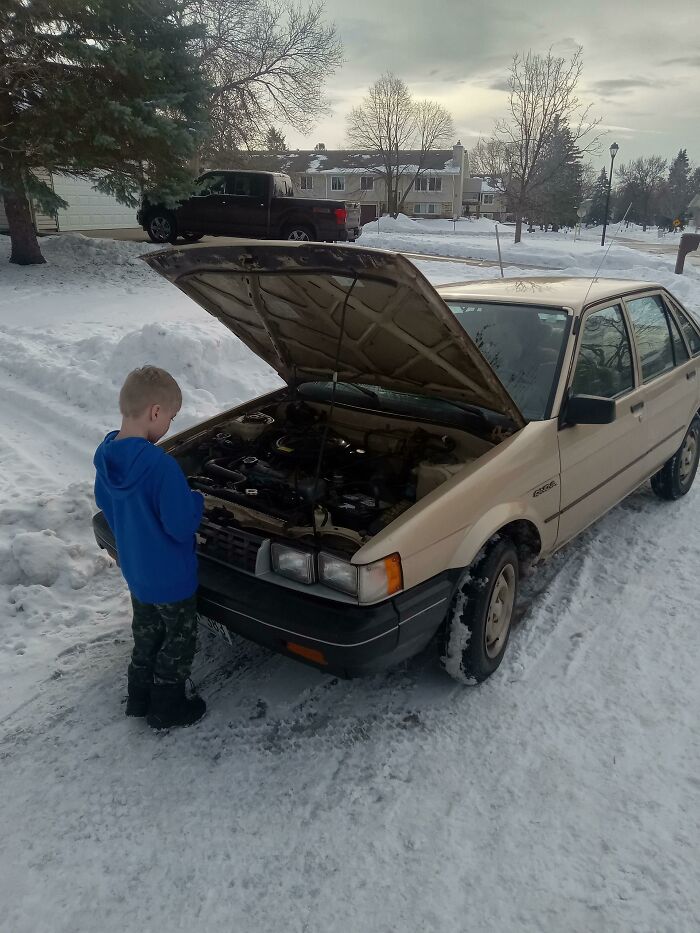 Child in blue jacket inspecting car engine with hood open on snowy street, capturing wholesome kind neighbors moment.