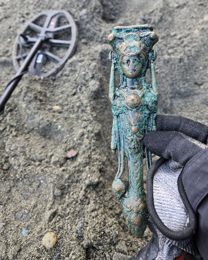 Gloved hand holding a detailed greenish metal statue found at the beach with a metal detector lying on the sand nearby.