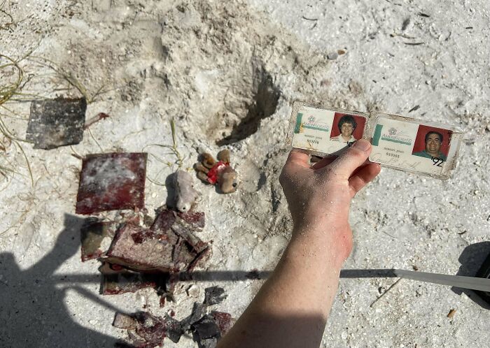 Hand holding old identification cards found among bizarre items on sandy beach, including worn toys and debris.