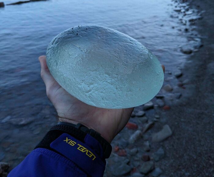 Hand holding a large translucent object found at the beach, with water and rocky shore in the background.