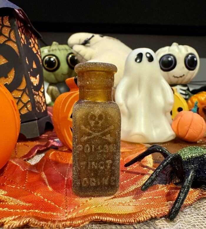 Frosted poison bottle with skull and crossbones among Halloween-themed objects and decorations.