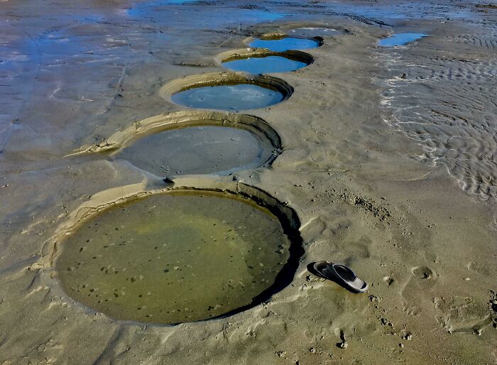 Series of large, unusual circular pools filled with water on a sandy beach with a single sandal nearby.