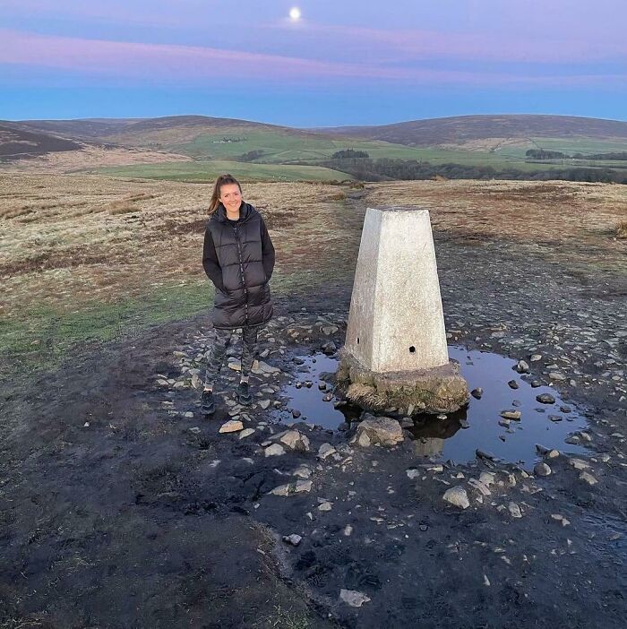 Woman wearing camo pants blending with surroundings standing near a stone marker in an open landscape matching surroundings.