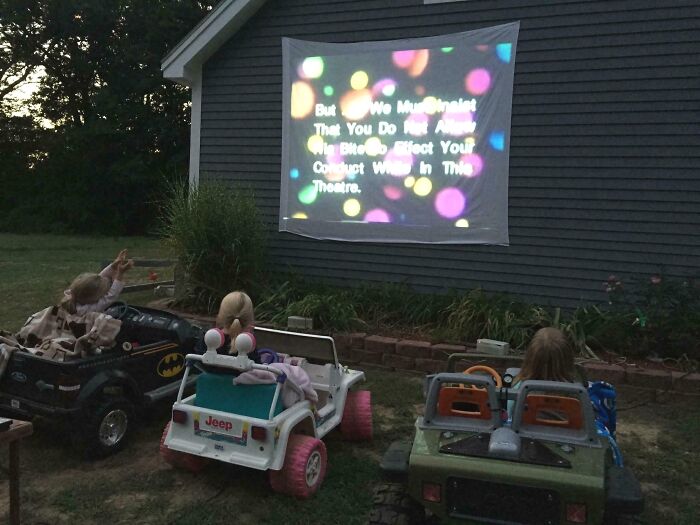 Three children in toy cars watching a movie projected on a house wall, showcasing wholesome kind neighbors.