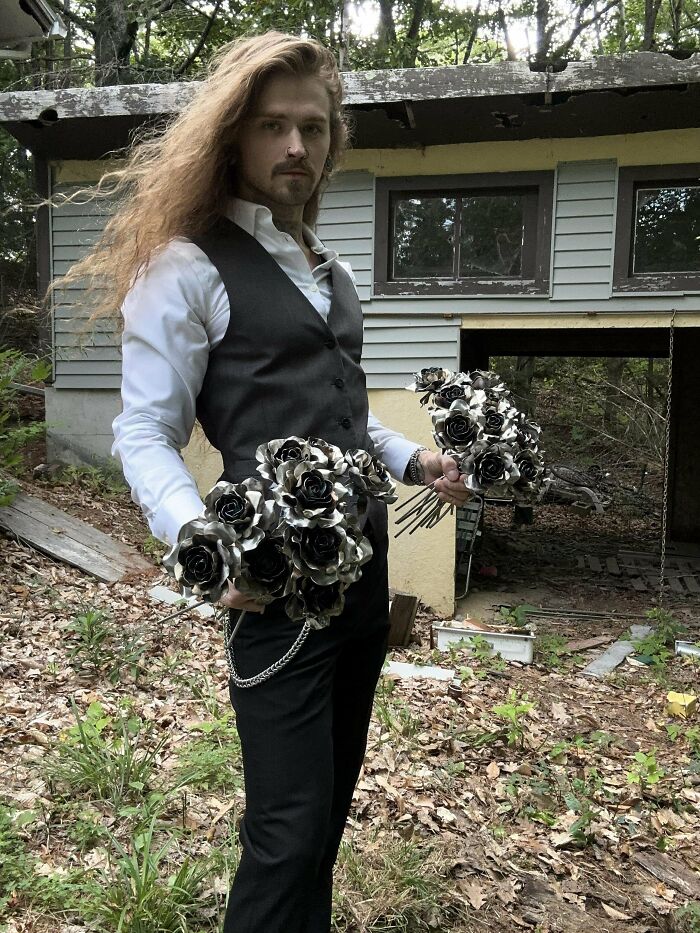 Man with fierceflow beautiful hair wearing a vest and holding metallic black and silver rose bouquets outdoors.