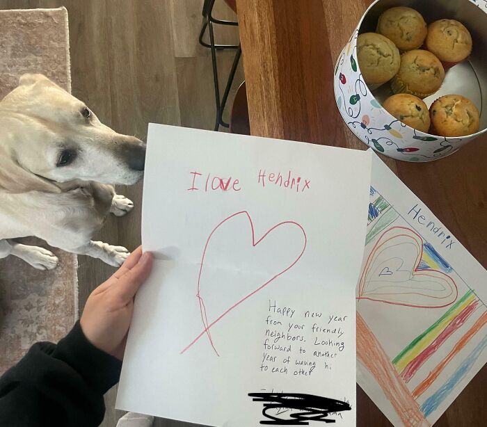 A person holding a handwritten note with a heart, a dog nearby, and muffins on a table, showing wholesome kind neighbors.