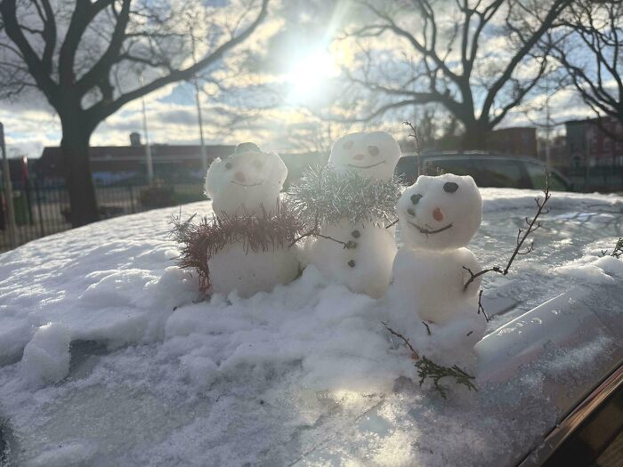 Three small snowmen with scarves and stick arms on a snowy car roof, capturing wholesome kind neighbors spirit.