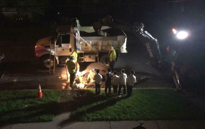 Neighbors gathered at night watching workers repair a street with construction equipment and bright lights.