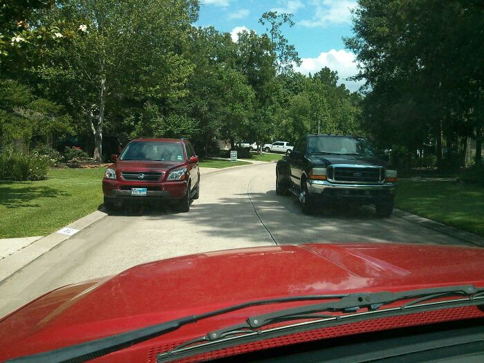Red car obstructed by two vehicles parked selfishly on both sides of a narrow suburban street blocking traffic flow.
