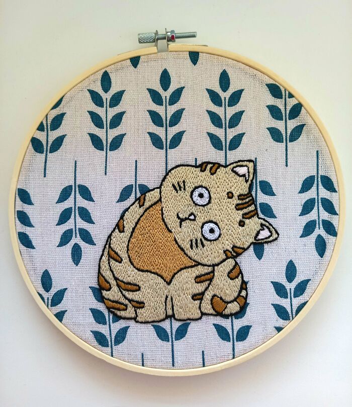 An adorable cat embroidery creation with wide eyes on a fabric with a teal leaf pattern.