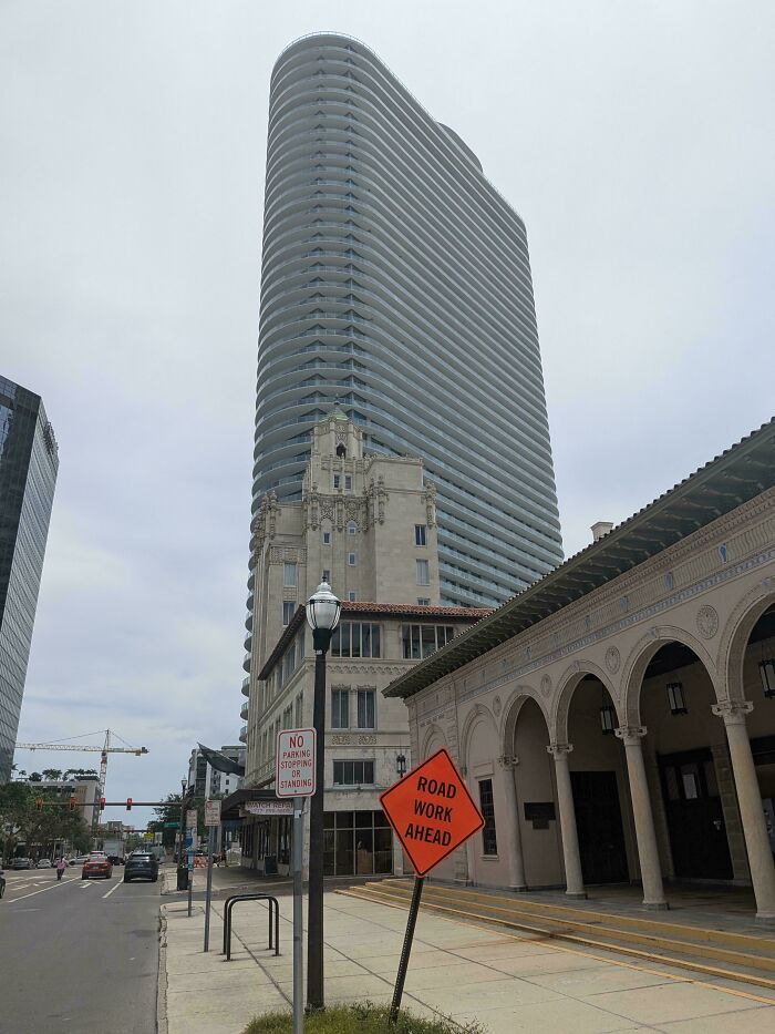 Modern high-rise building towering behind old architectural structures on a city street with construction signs visible.