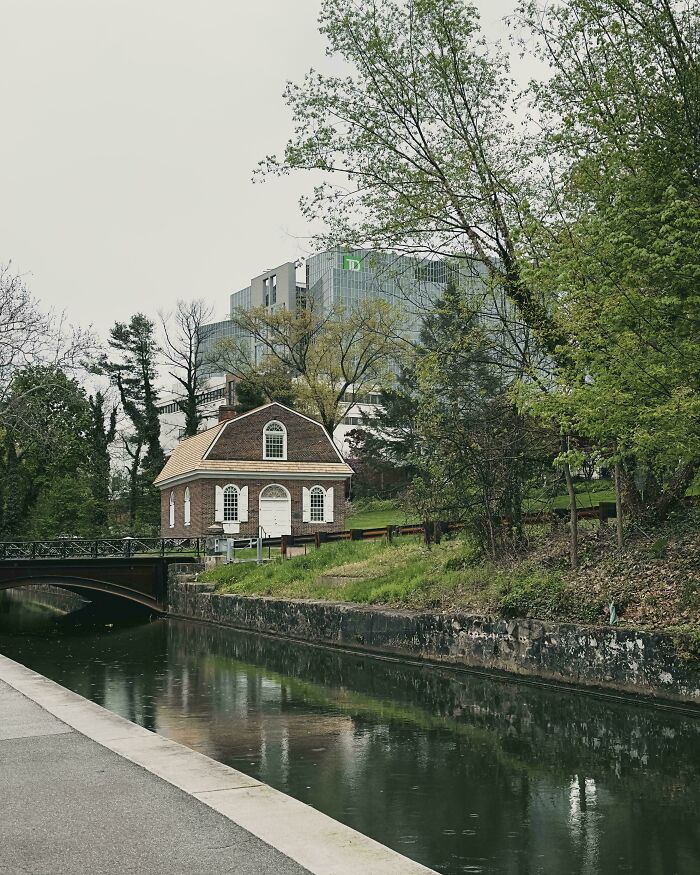 Small old brick house by a canal contrasted with modern glass office buildings behind, surrounded by trees.