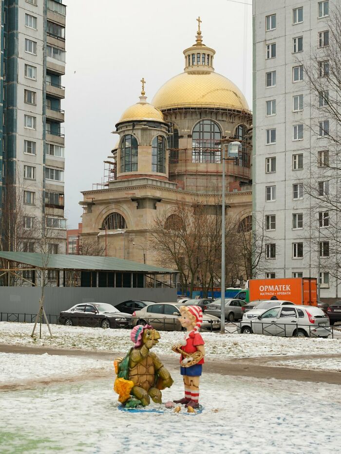 Statues of a cartoon boy and turtle in a snowy park with old golden-domed church and modern apartment buildings nearby.