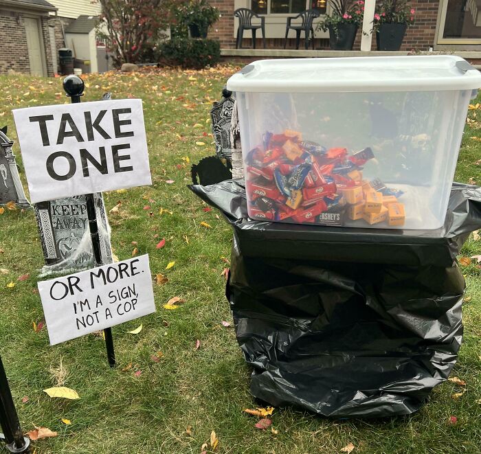 Plastic bin of candy with humorous signs on lawn inviting to take one or more, showcasing random notes and messages.