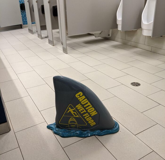 Shark fin-shaped caution wet floor sign on tiled restroom floor with humorous design to prevent slipping.
