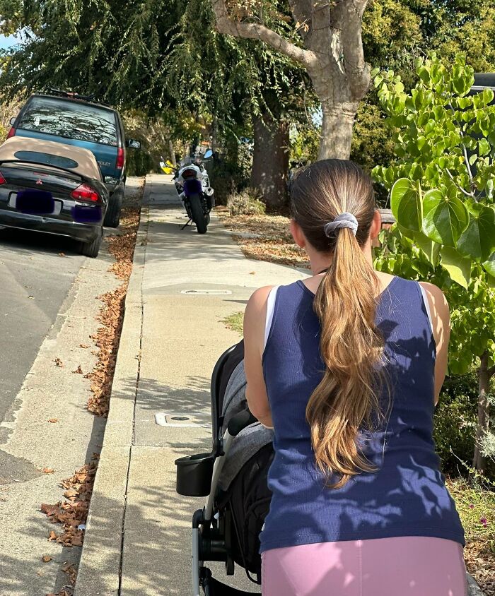 Woman in a blue top pushing a stroller on a sidewalk blocked by a motorcycle and parked cars showing selfish people causing inconvenience.