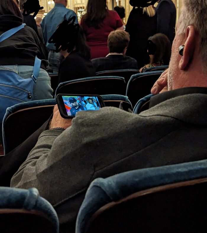 Man in a crowded setting watching a video on his phone, showing selfish people causing inconvenience to others.