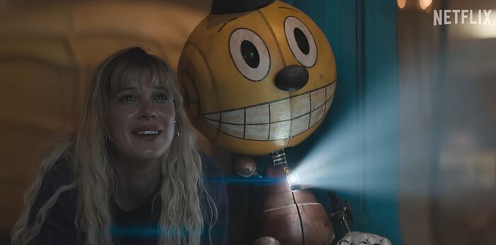 Woman with blonde hair sitting next to a large smiling robot toy, scene from a Netflix movie that turned out to be a letdown.