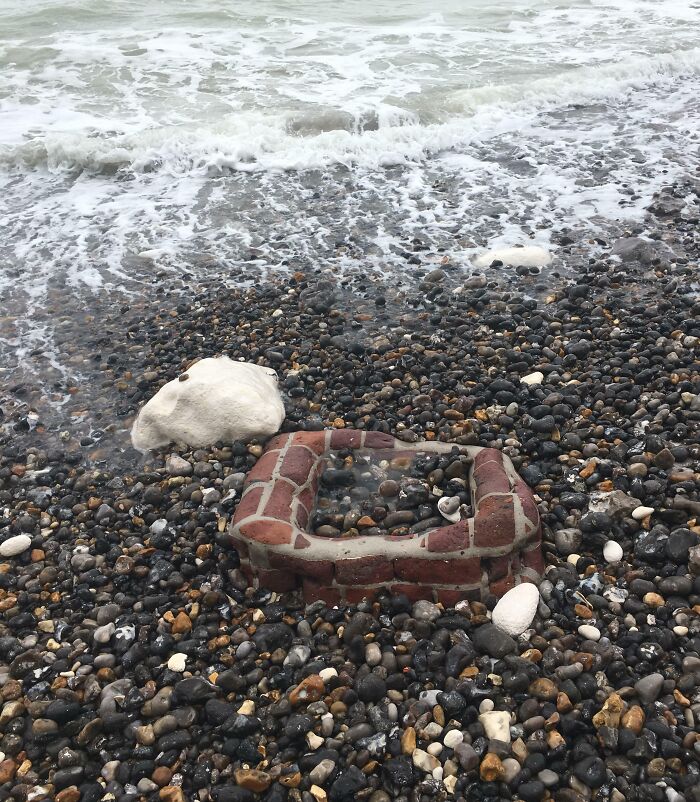 Brick structure partially buried among pebbles on a beach shore with waves in the background, an interesting beach find.