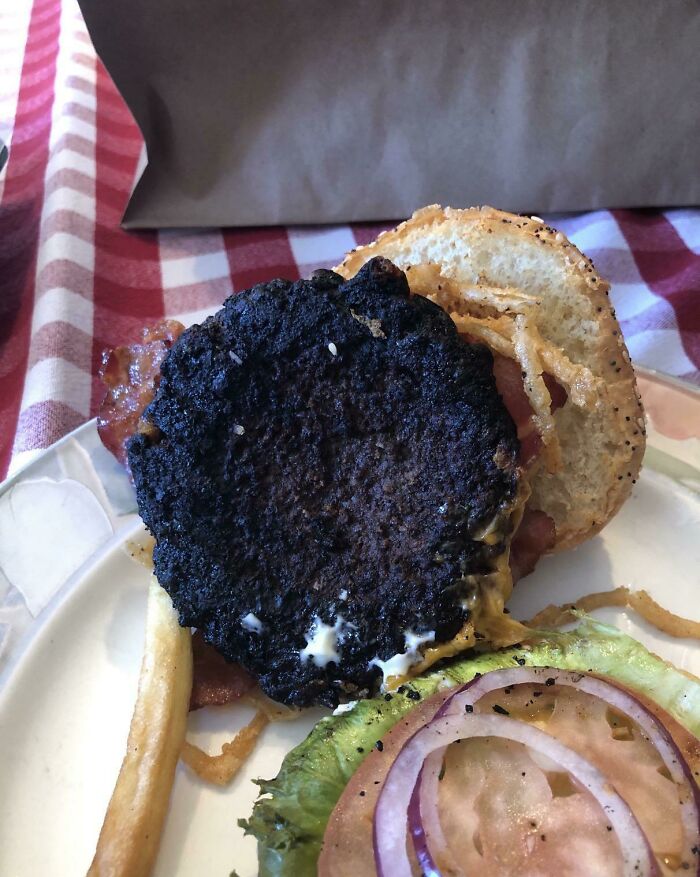 Burnt burger patty on sandwich with lettuce, tomato, onion, and fries, showcasing restaurant workers dealing with unusual orders.