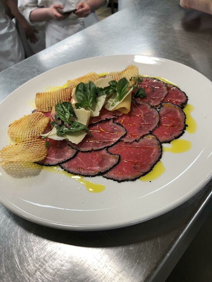Plate of thinly sliced beef carpaccio with greens, shaved cheese, and crispy waffle-cut potato chips in a restaurant kitchen.