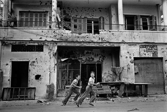 Two boys running past a heavily damaged building with bullet holes in this weird history photo showing war impact.