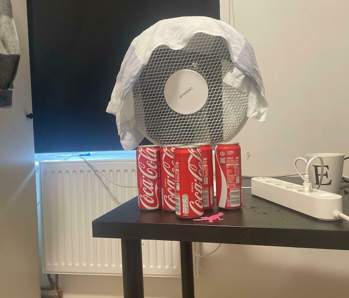 Small fan elevated on Coca-Cola cans with a cloth draped over it, demonstrating a genius life hack for cooling.