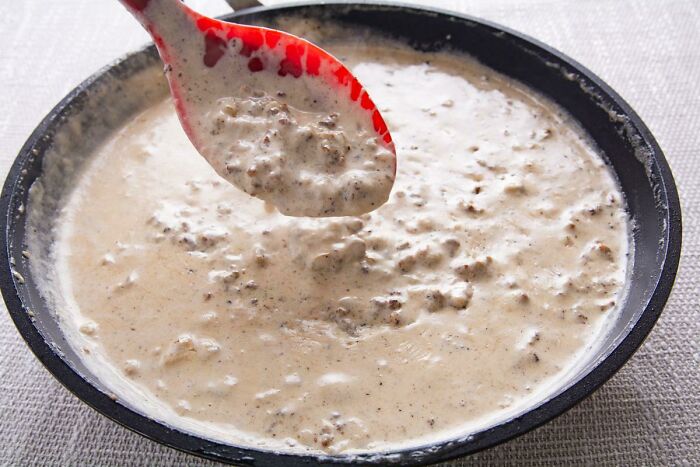 Creamy sausage gravy in a skillet with a red spoon, a classic comfort food that restaurant workers often serve.