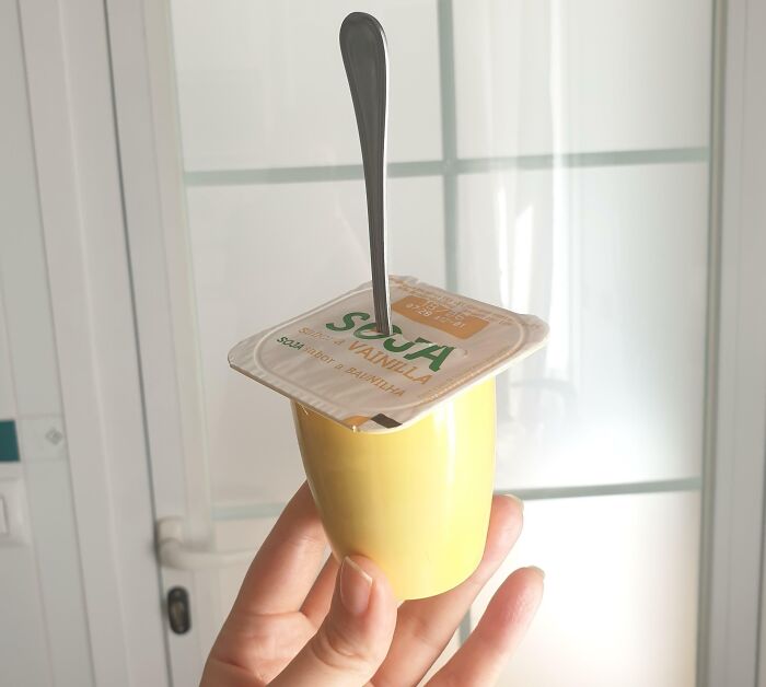 Hand holding a yellow cup with a vanilla soy yogurt lid pierced by a spoon demonstrating a genius life hack.