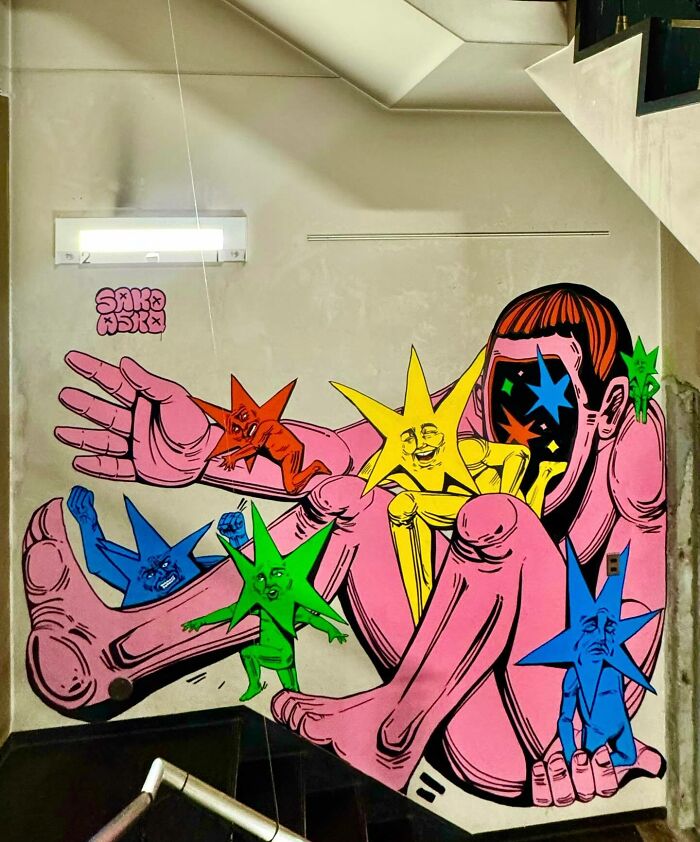 Mural by Colombian artist SakoAsko featuring vibrant humanoid stars interacting with a large pink figure on a wall.