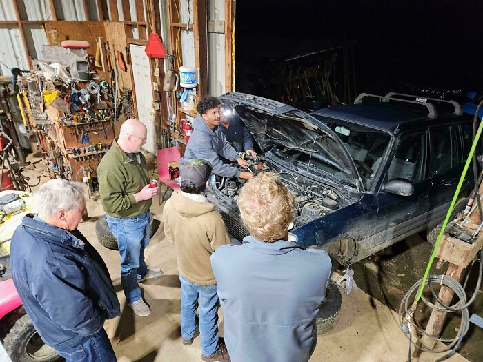 Car mechanics gathered around an open engine, taking pictures to show unusual car repairs and issues.