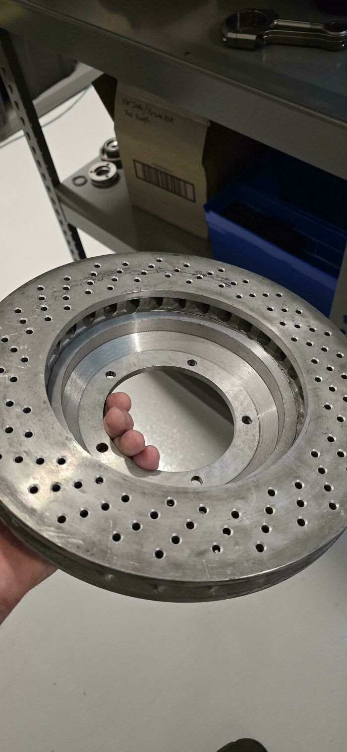 Car mechanic holding a large drilled brake rotor, showcasing the unusual size and design in the workshop setting.