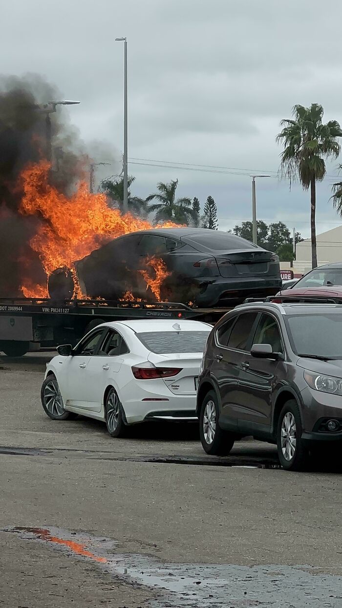 Black car on fire on a tow truck with thick smoke rising while other vehicles are parked nearby in a mechanic area