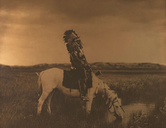 Native American in traditional dress riding a white horse drinking water in a grassland, a significant 20th century image.