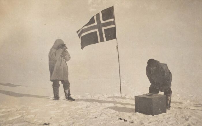 Two explorers in snowy terrain beside a Norwegian flag, capturing a moment of 20th century triumph and tragedy.