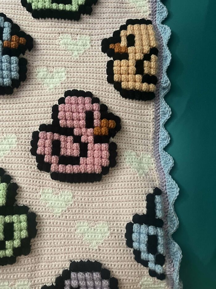 Crocheted blanket featuring pixelated colorful ducks with heart patterns and a scalloped edge on a green background.