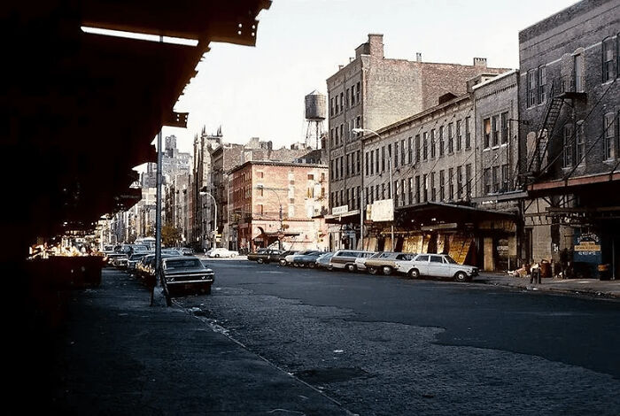 Urban hell scene showing rundown buildings, empty sidewalks, and parked cars in a neglected city neighborhood.