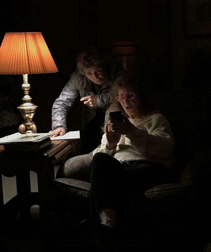 Two women illuminated by a lamp, one sitting and looking at a phone, the other leaning over in a scene like a Renaissance masterpiece.