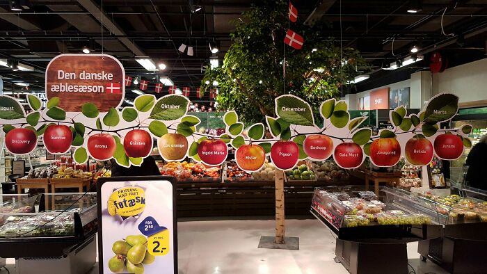 Display of Danish apple varieties with flags and seasonal labels in a grocery store, highlighting Danish culture and produce.
