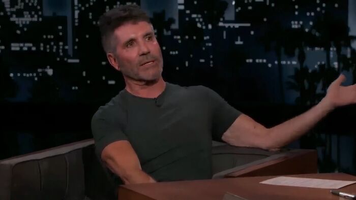 Man in a dark t-shirt gesturing while discussing work and life on a late-night talk show set with city lights background.