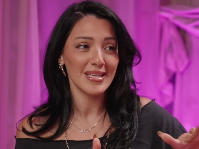 Woman with black hair gesturing while discussing work and life, highlighting celebrity entitlement in a purple-lit setting.