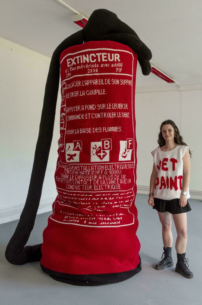 Large crocheted fire extinguisher sculpture next to a woman wearing a knitted vest, showcasing crocheting art and craftsmanship.