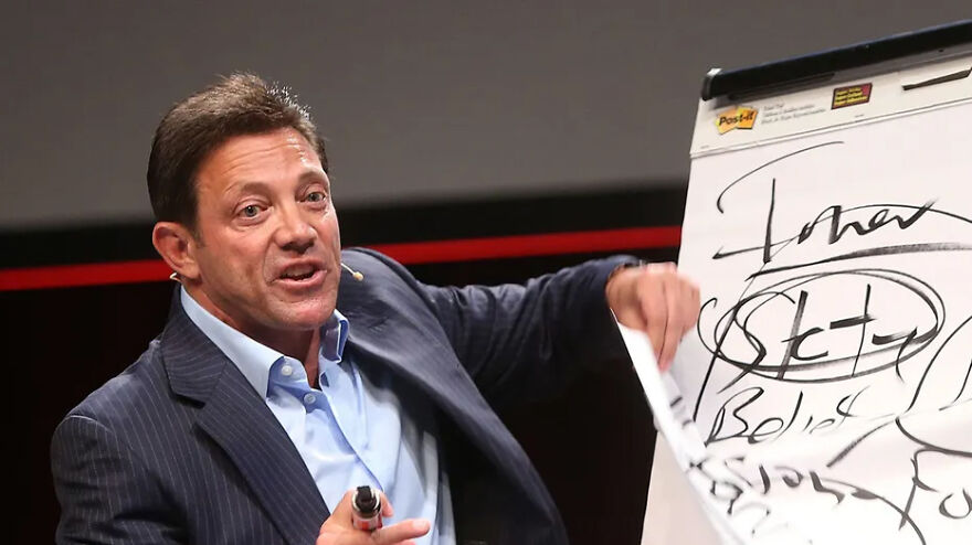 Jordan Belfort in a suit speaking and writing on a flip chart, illustrating financial concepts about millions owed.